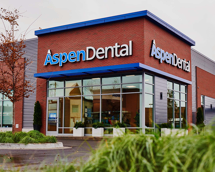 Exterior of an Aspen Dental clinic with a red brick facade, large glass windows, and blue trim, featuring prominent “Aspen Dental” signage above the entrance.