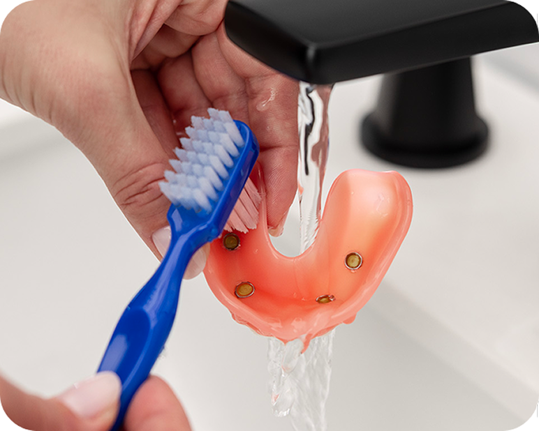 Alt text:
Person cleaning an implant-supported denture with a toothbrush under running water.
