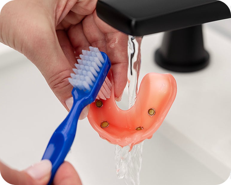 Person cleaning an implant-supported denture with a toothbrush under running water.