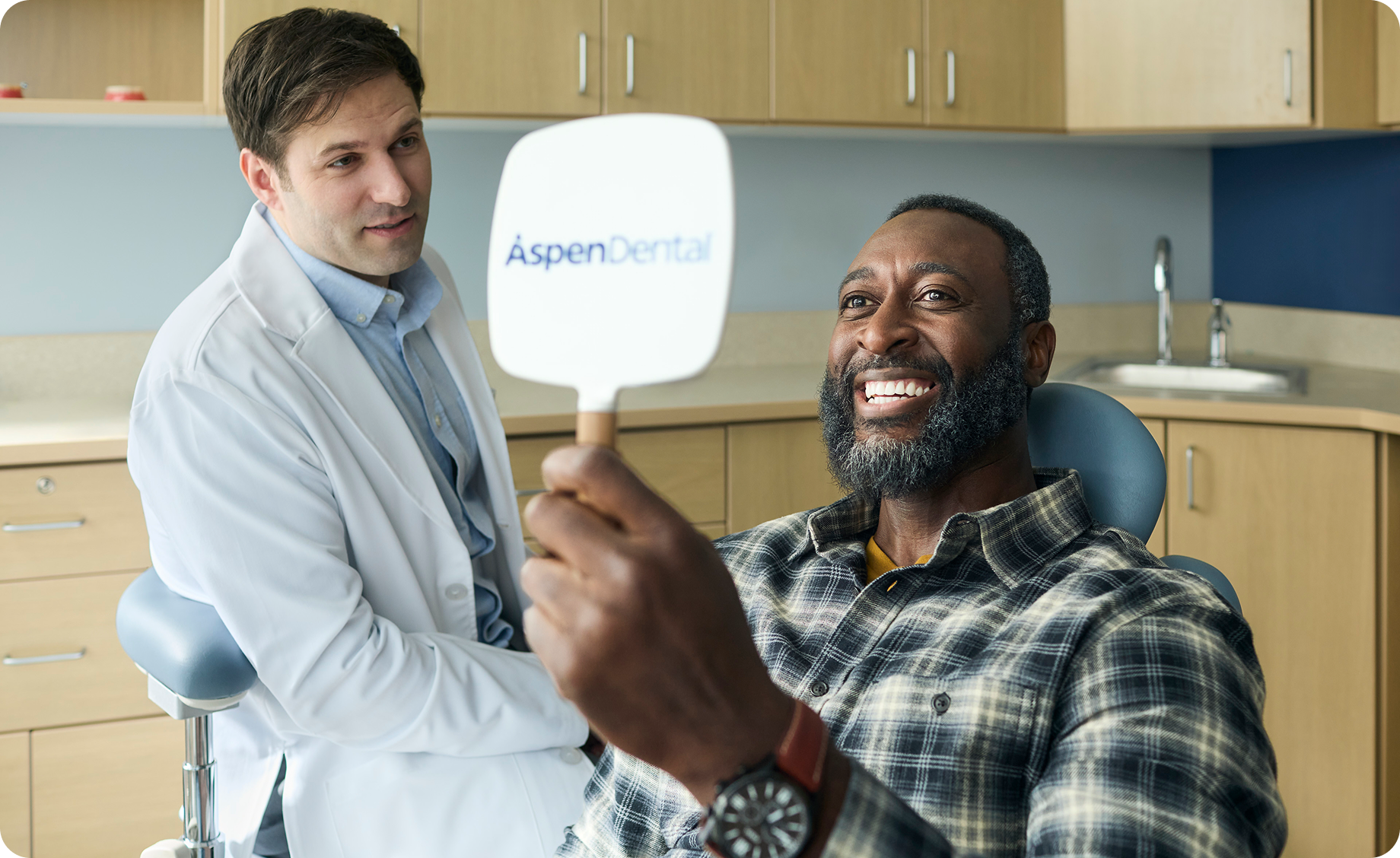 Smiling adult man holding an Aspen Dental mirror and viewing his smile during a dental visit, with a dentist in the background.