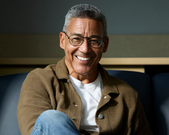 Alt text: Smiling adult man wearing glasses and a casual jacket seated indoors with a relaxed, confident expression.