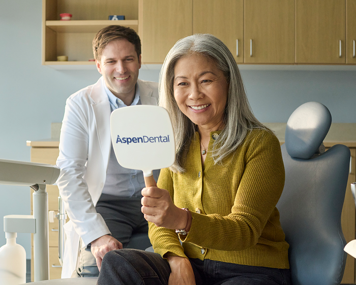 Smiling older woman holding a mirror and viewing her smile during a dental visit, with a dentist in the background.