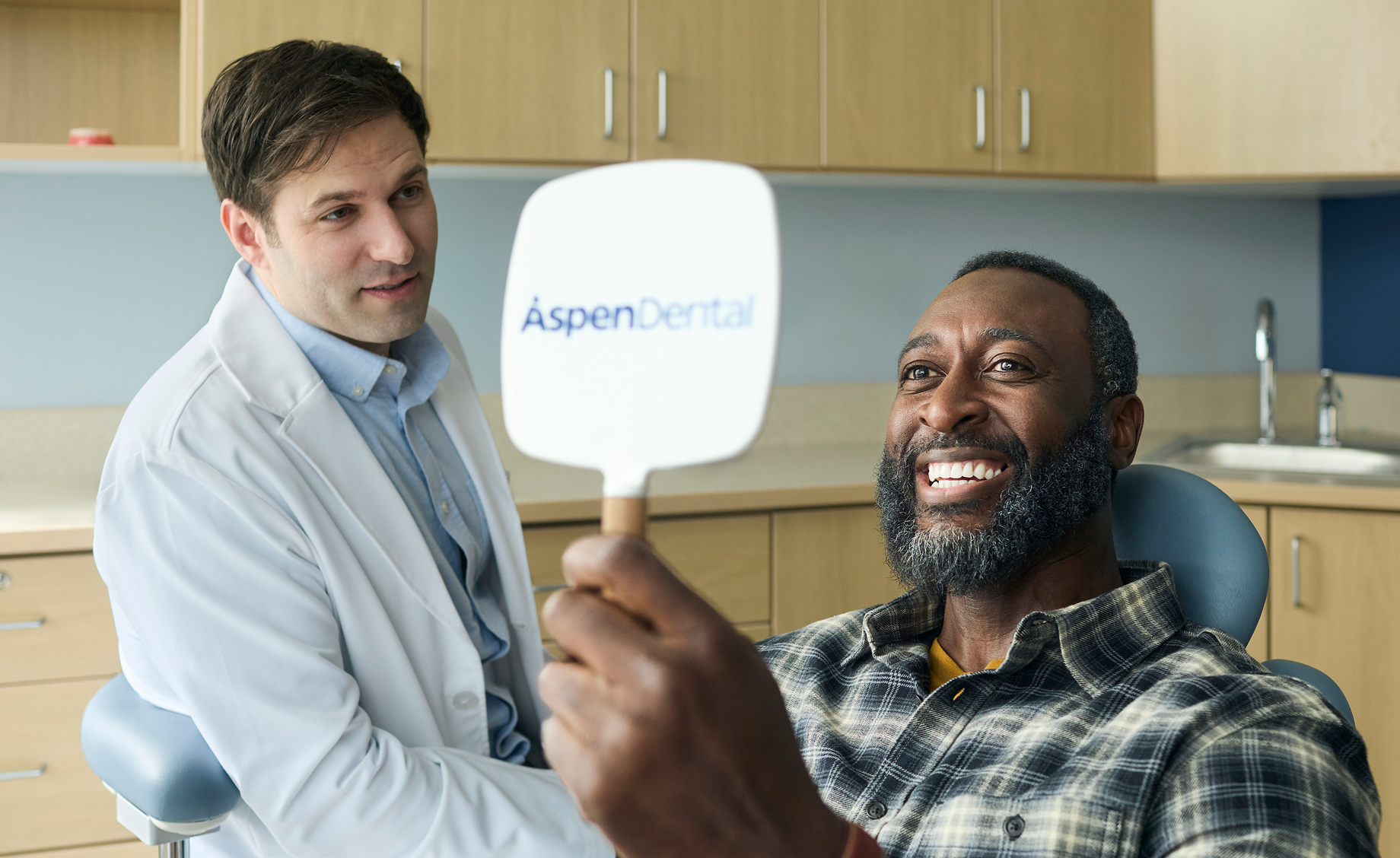 Smiling adult man holding an Aspen Dental mirror and viewing his smile during a dental visit, with a dentist in the background.