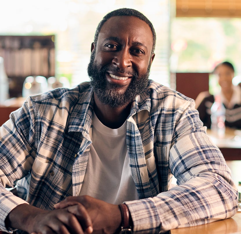  A smiling male patient sits at a table in a relaxed café setting, looking toward the camera.
