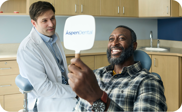 Alt text:
Patient holding an Aspen Dental mirror and smiling during a dental visit with a dentist nearby.