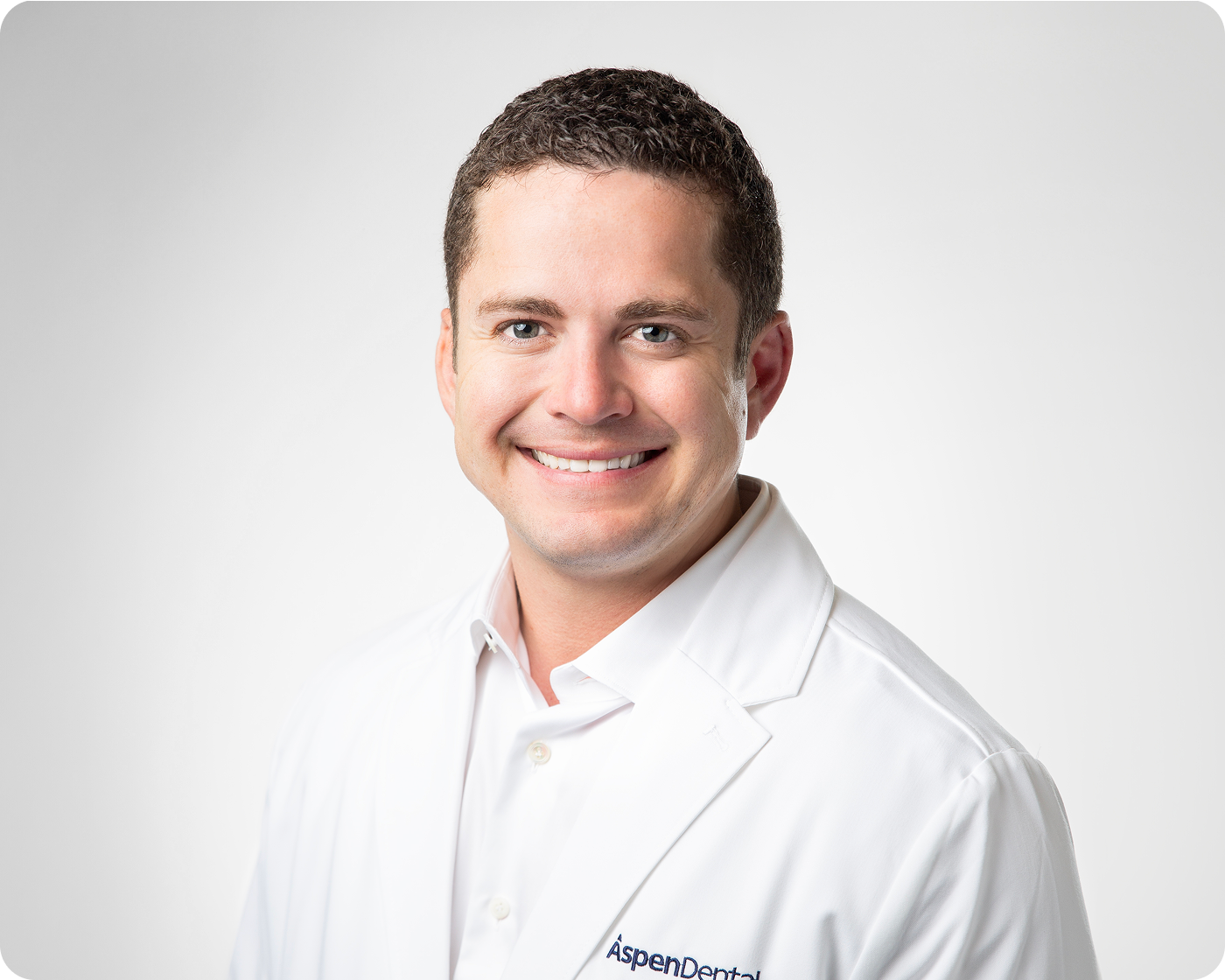 Professional headshot of a smiling male dental provider wearing a white coat against a gray background