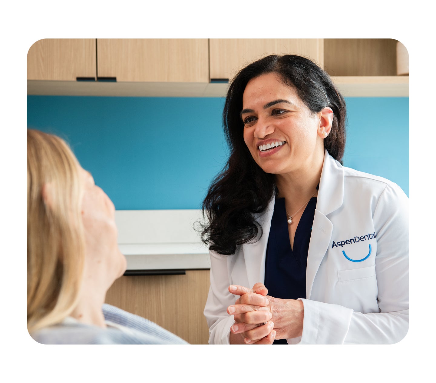 An Aspen Dental dentist smiles warmly while speaking with a seated patient during a one-on-one consultation in a modern dental office.