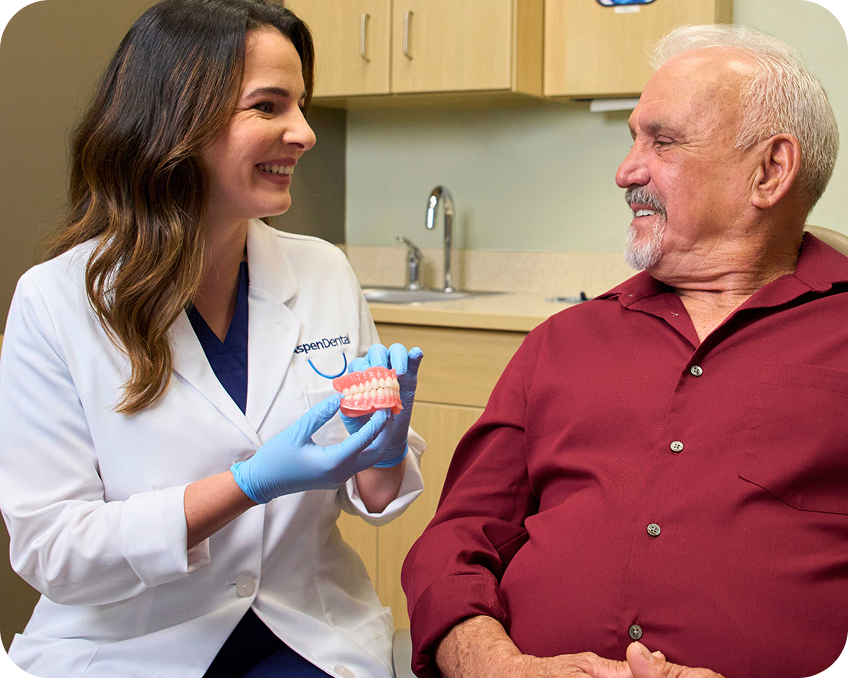 A dental professional in a white coat and blue gloves showing a teeth model to an elderly patient in a dental office
