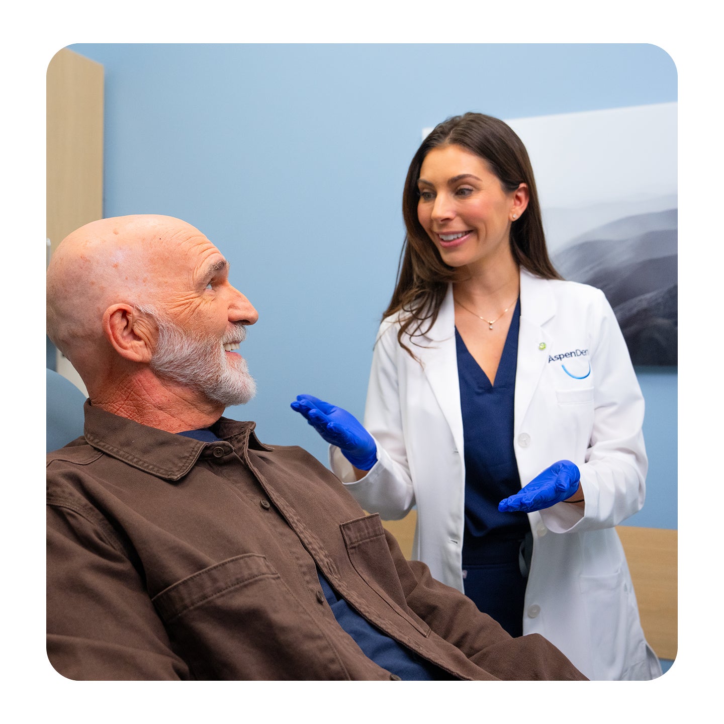 An Aspen Dental dentist wearing a white coat and blue gloves speaks with a male patient seated in a dental exam chair during a consultation.