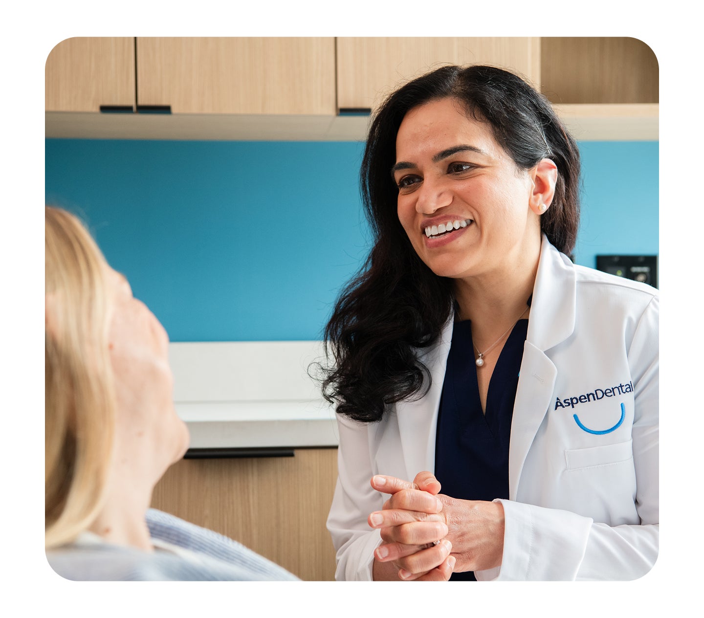 A smiling Aspen Dental dentist in a white lab coat with the Aspen Dental logo, speaking to a patient reclining in a dental chair.
