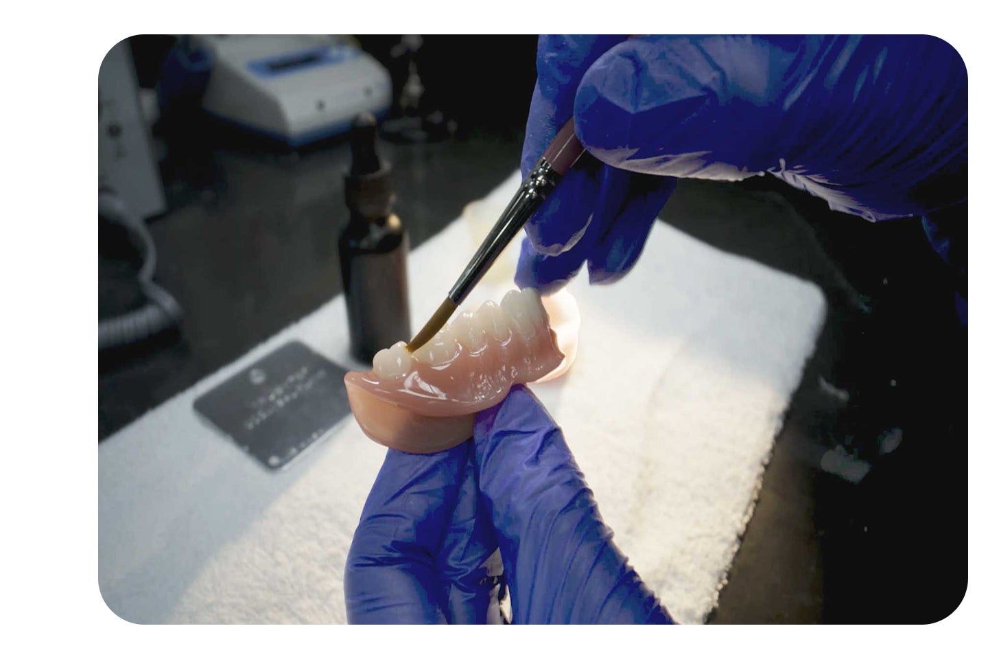 Gloved hands apply material to the underside of a denture using a brush, illustrating precise lab work involved in a professional denture reline at Aspen Dental.
