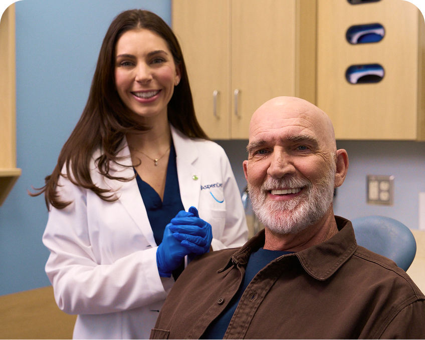 An Aspen Dental dentist in a white coat smiles beside a male patient seated in the exam chair, both looking happy after dental treatment.
