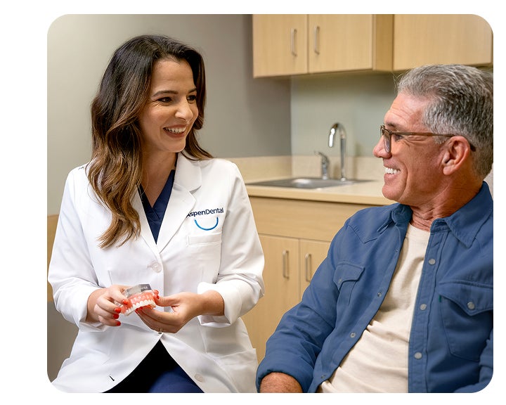 A woman in a dental coat holds a denture while talking to an older man