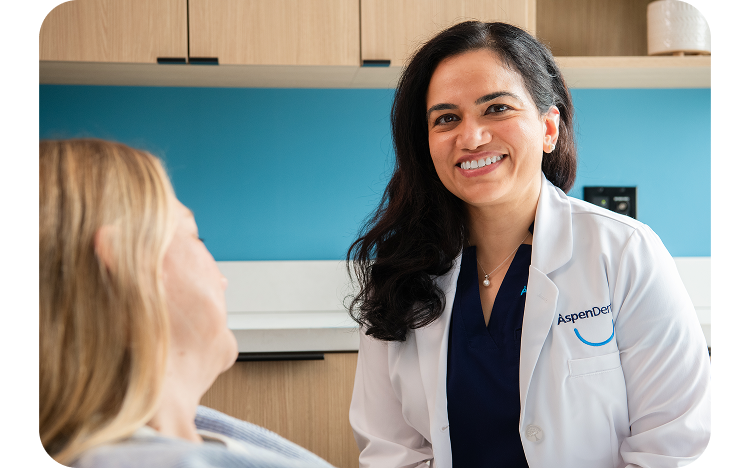 Aspen Dental dentist in a white coat smiling confidently at a female patient during a consultation in an exam room.