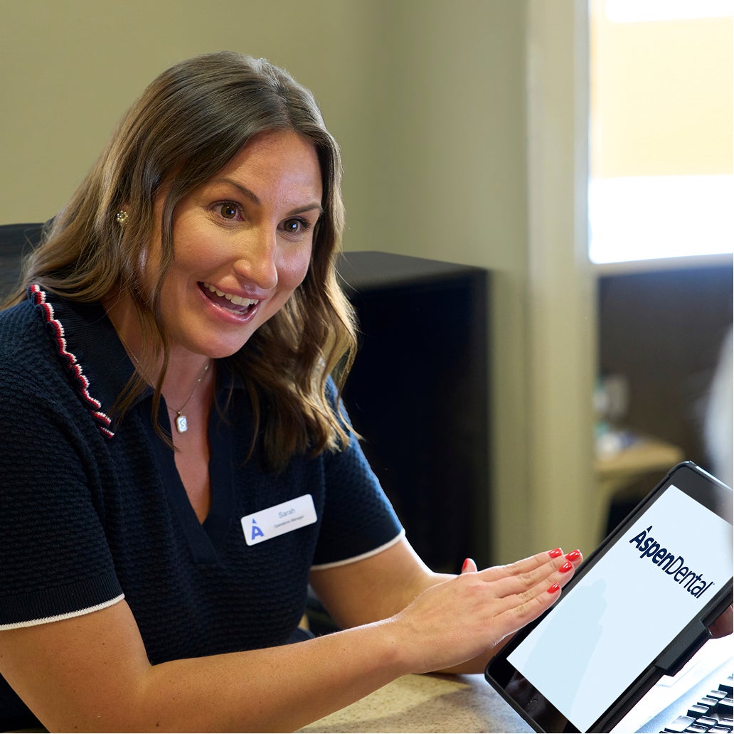 An Aspen Dental team member smiles while reviewing information on a tablet with a patient, explaining treatment or payment options during a consultation.