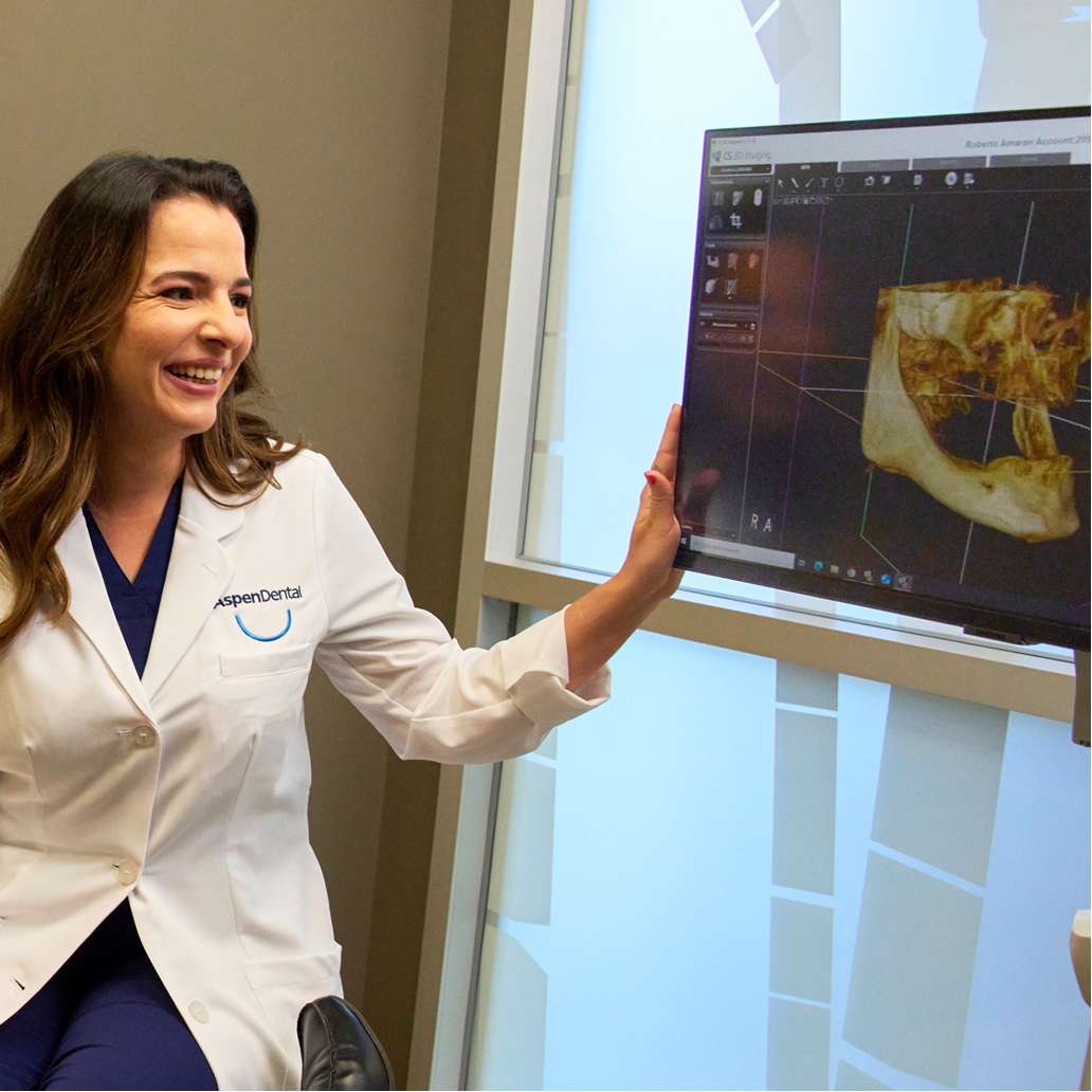 An Aspen Dental dentist in a white coat smiles while reviewing a 3D dental X-ray image displayed on a monitor during a patient consultation.