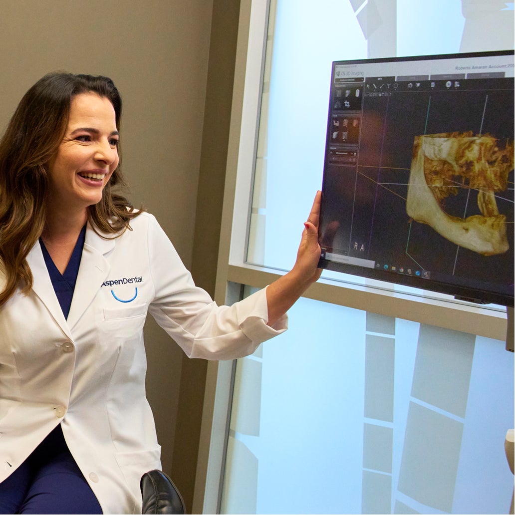 An Aspen Dental dentist in a white coat smiles while reviewing a 3D dental X-ray image displayed on a monitor during a patient consultation.