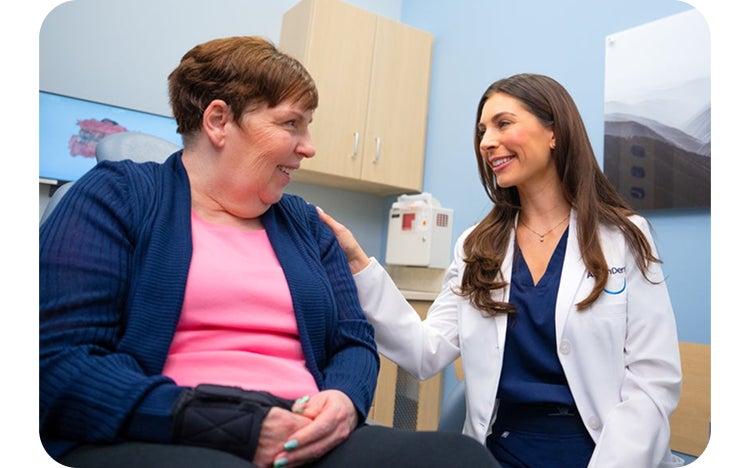 An Aspen Dental dentist in a white coat smiles while gently reassuring a seated female patient during a consultation in a dental exam room.