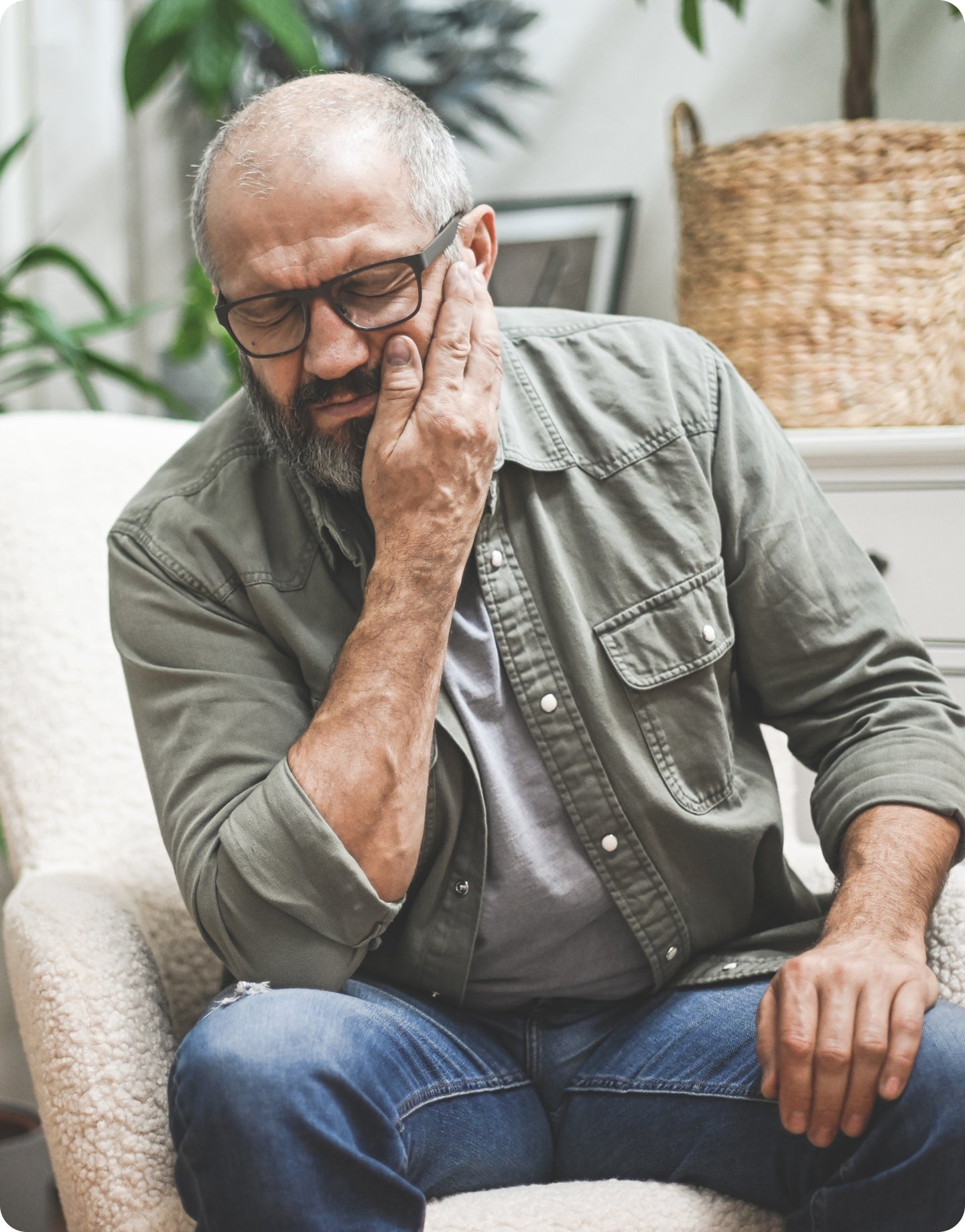 Man sitting on a couch holding his cheek with a pained expression, suggesting toothache pain.