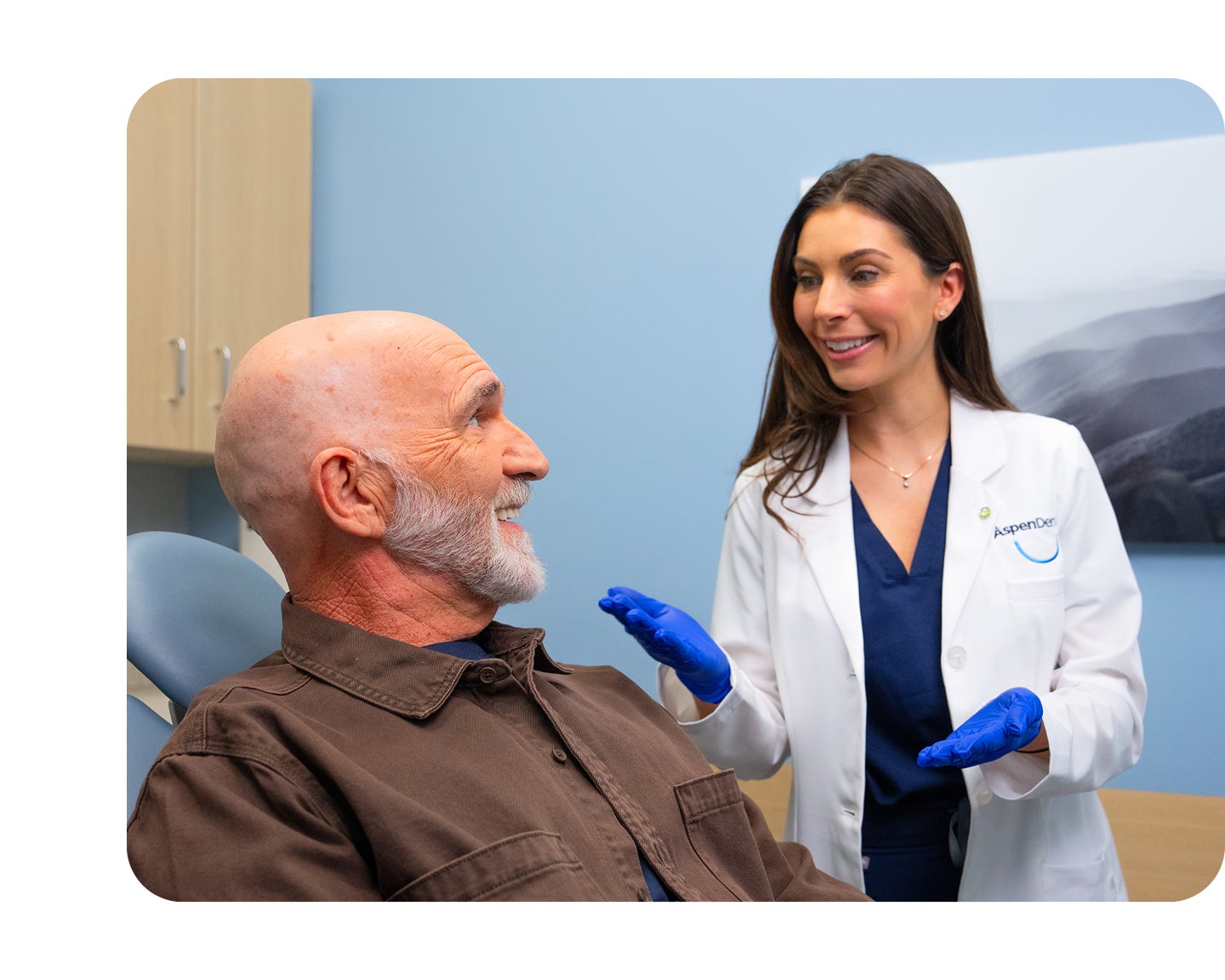 An Aspen Dental dentist wearing blue gloves speaks with a smiling male patient seated in a dental exam chair during a routine checkup.