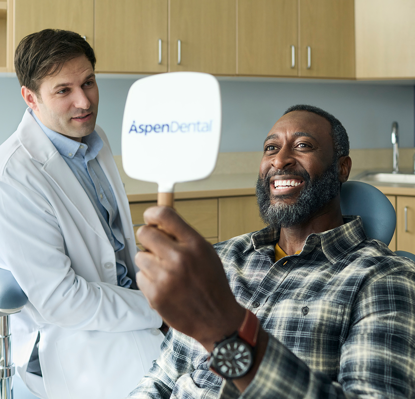 Smiling adult man holding an Aspen Dental mirror and viewing his smile during a dental visit, with a dentist nearby.