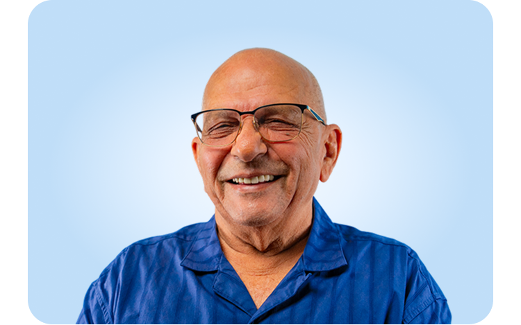 A close-up shot of a smiling senior man with glasses and a shaved head, wearing a blue shirt. He is laughing and looking directly at the camera.