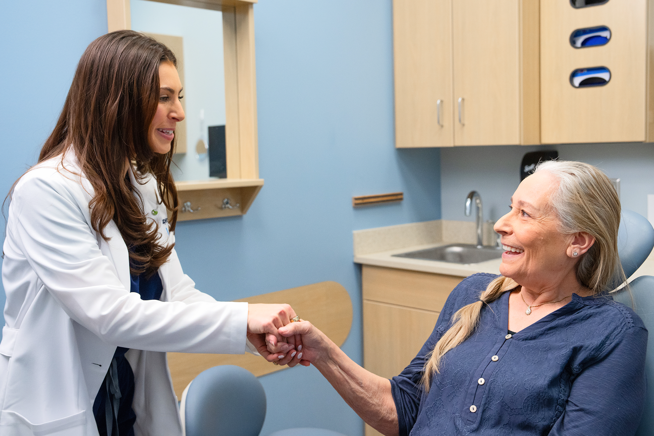 Two women are seated on a couch, engaged in conversation. One woman gestures with her hands while speaking, and the other listens attentively with her back to the camera.
