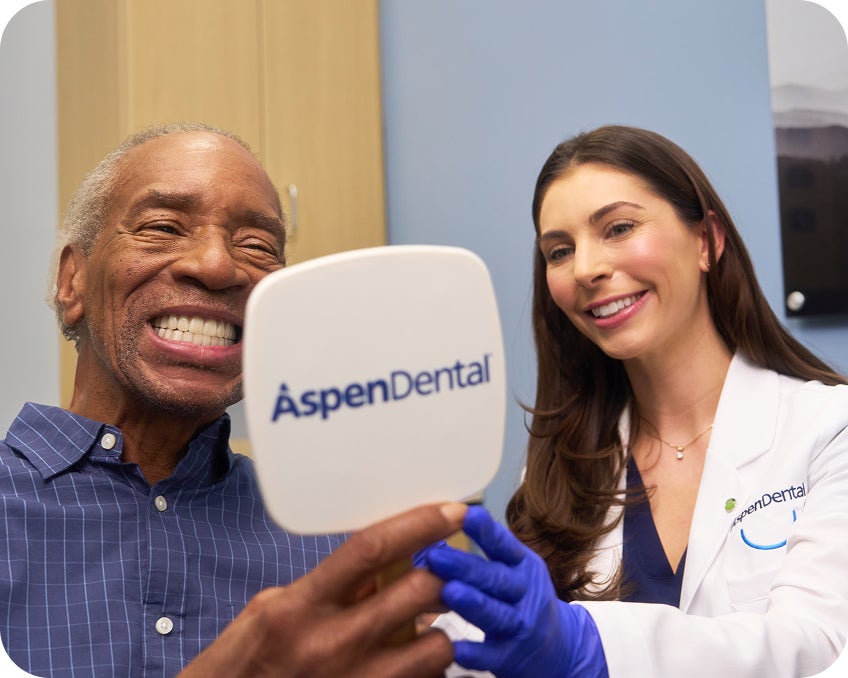 A male patient smiles while looking into an Aspen Dental mirror, with a dentist in a white coat smiling beside him during the appointment.
