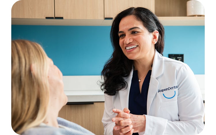 A smiling Aspen Dental dentist in a white lab coat with the Aspen Dental logo, speaking to a patient reclining in a dental chair.