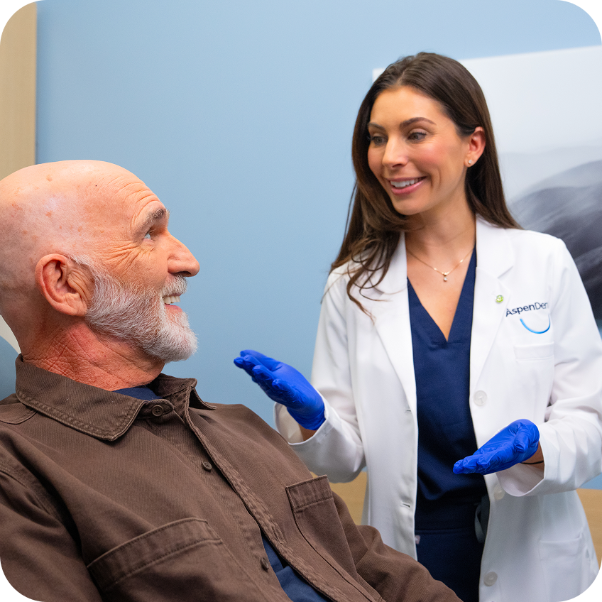 An Aspen Dental dentist in a white coat and blue gloves speaks with a smiling male patient seated in a dental exam chair during a consultation.
