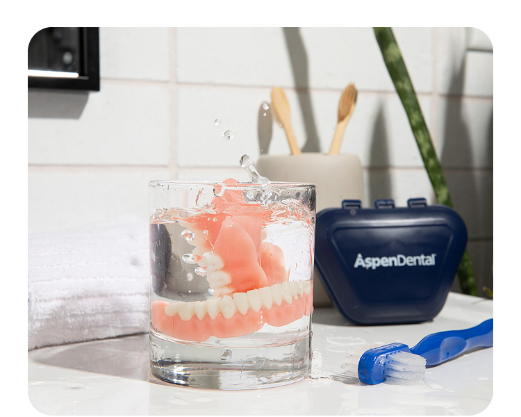 Dentures in a glass of water on a bathroom counter with a blue toothbrush, Aspen Dental container, wooden toothbrushes, and a white towel.