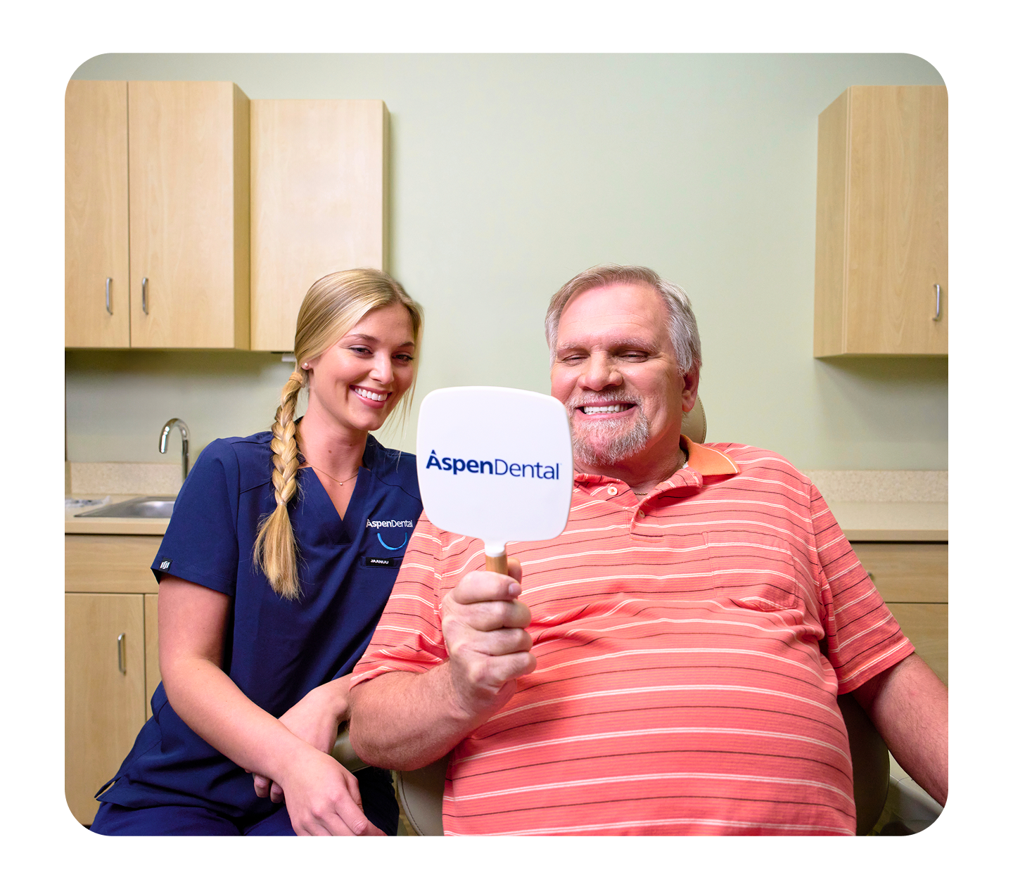 A smiling male patient holds an Aspen Dental mirror while seated in a dental exam chair, as a dental assistant in navy scrubs smiles beside him during the visit.