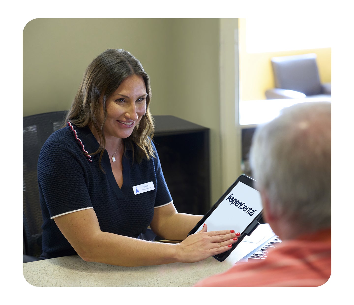 An Aspen Dental team member smiles while showing information on a tablet to a seated patient during a one-on-one consultation at the front desk.