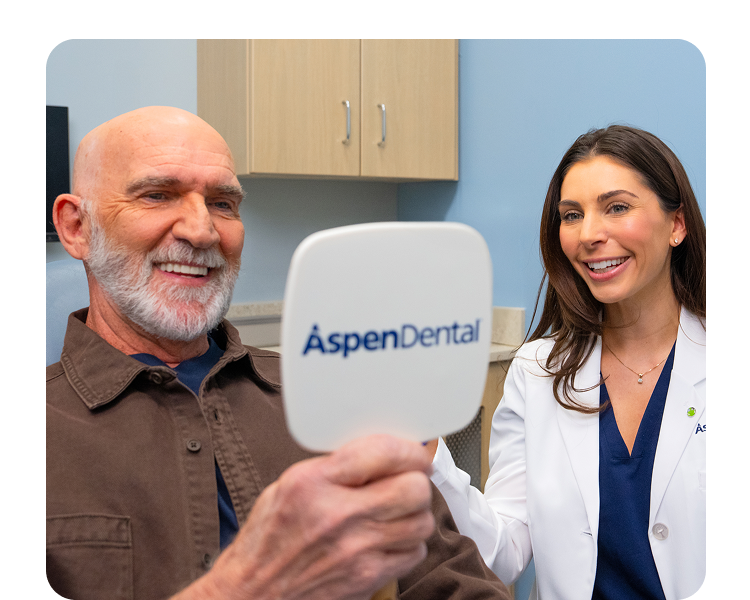  A happy Aspen Dental patient admires his smile in a handheld mirror after receiving dental implants as a smiling Aspen Dental dentist stands next to him with her hand on the patients shoulder. The image conveys confidence symbolizing the benefits of dental implants.