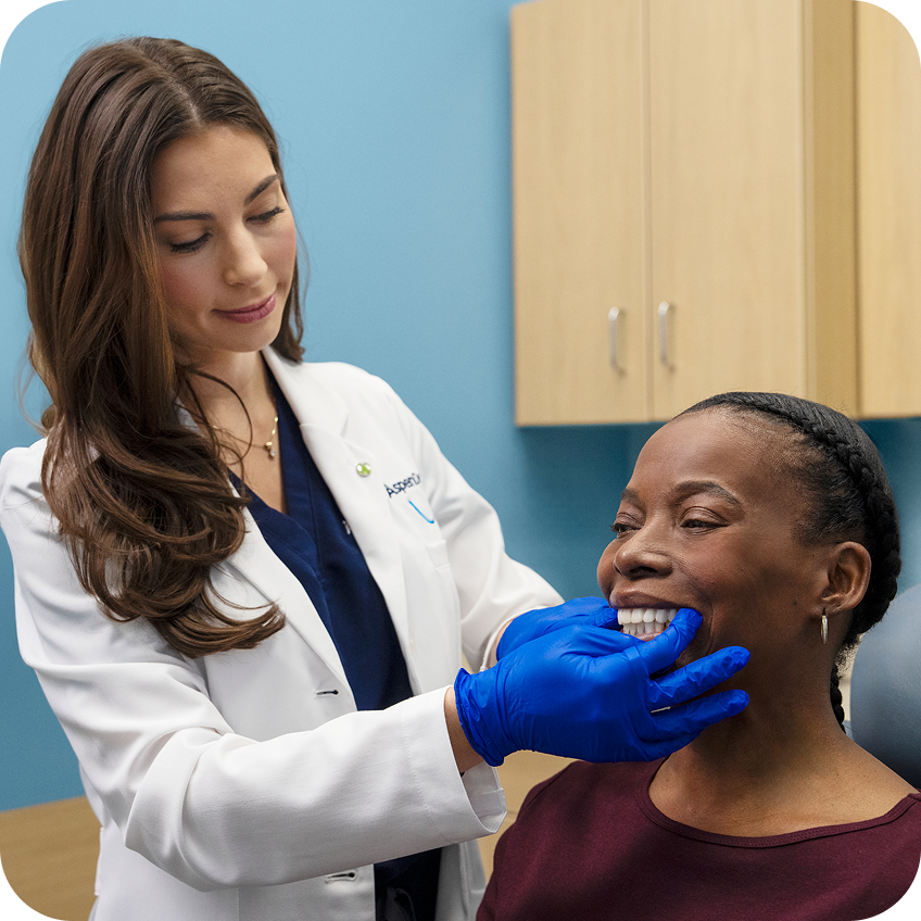 An Aspen Dental dentist wearing blue gloves gently examines a smiling patient’s teeth during an in-chair dental exam in a modern exam room.
