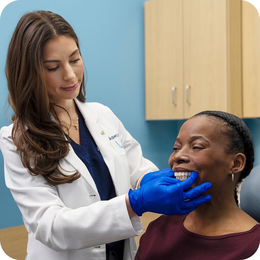 An Aspen Dental dentist wearing blue gloves gently examines a smiling patient’s teeth during an in-chair dental exam in a modern exam room.