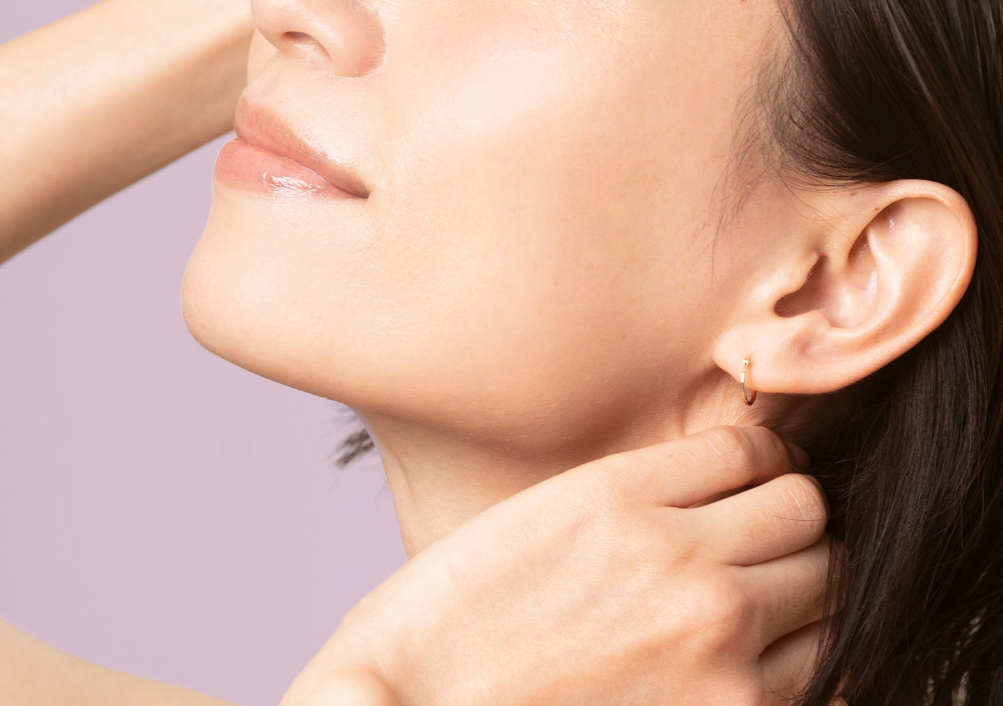 A closeup of a woman with smooth, radiant skin touching her ear with her hand, set against a lavender background
