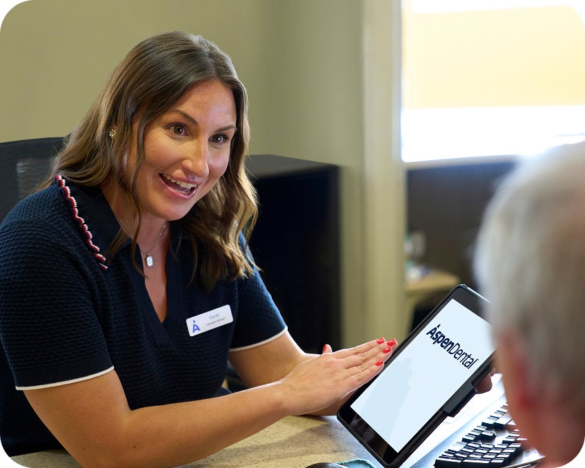 An Aspen Dental team member smiles while showing a patient information on a tablet during a consultation at the front desk.
