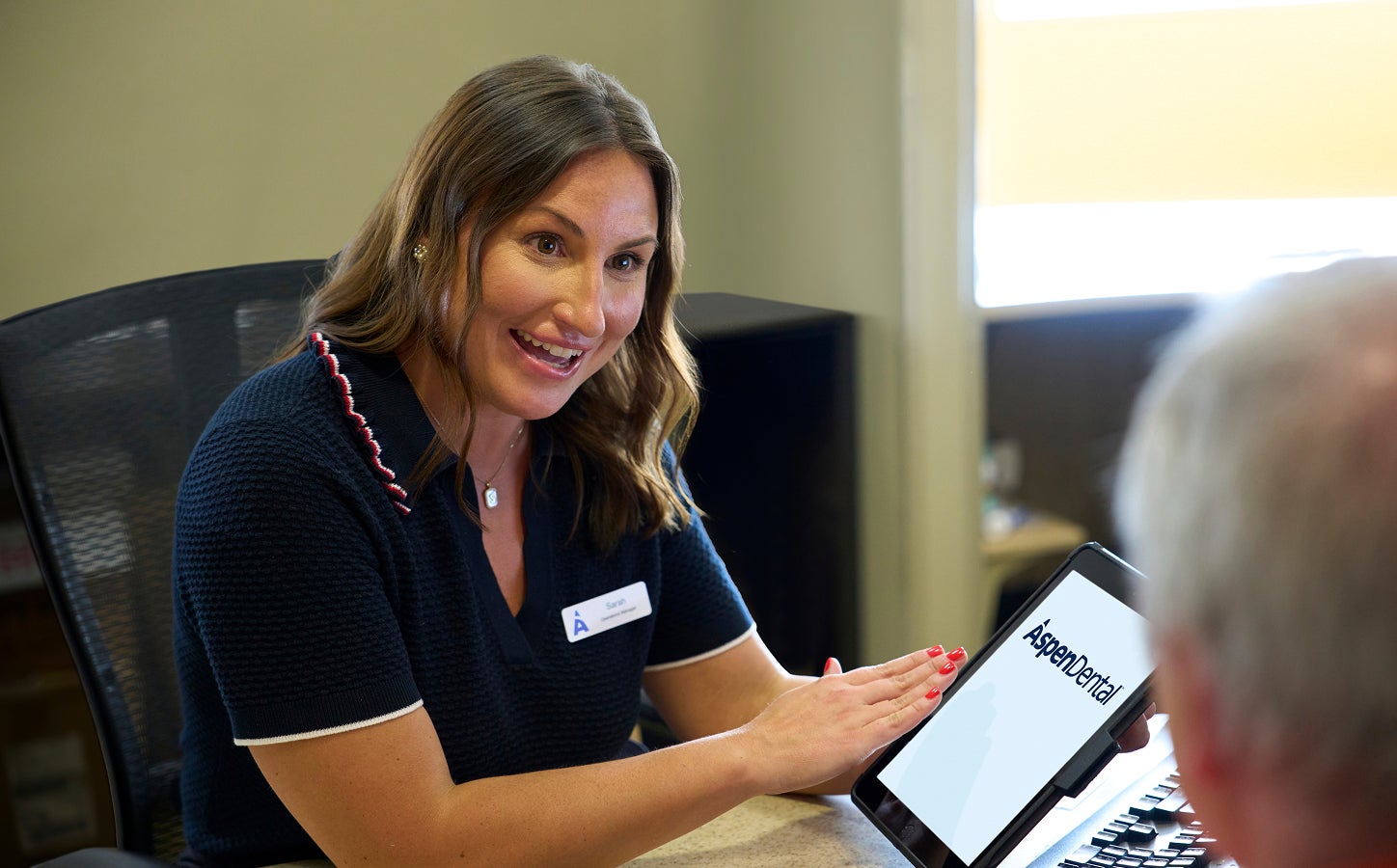 An Aspen Dental team member smiles while reviewing information on a tablet with a patient across the desk during a consultation.