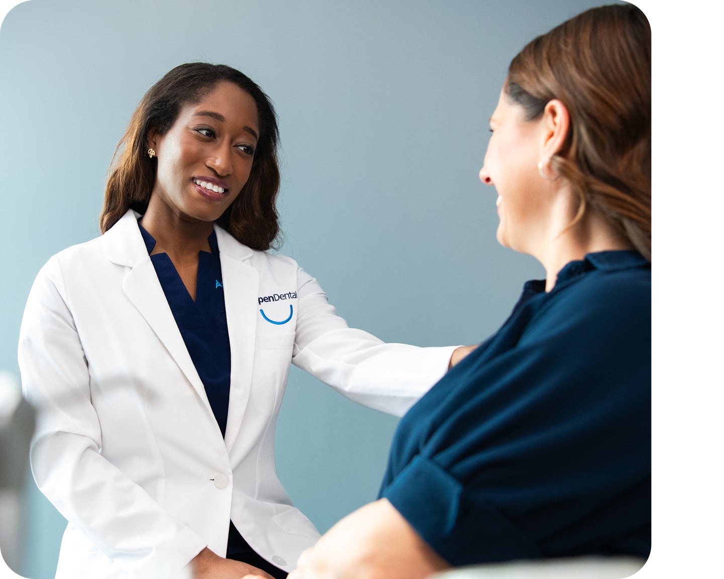 An Aspen Dental dentist in a white coat gently speaks with a seated patient, offering reassurance during a consultation in a modern exam room.