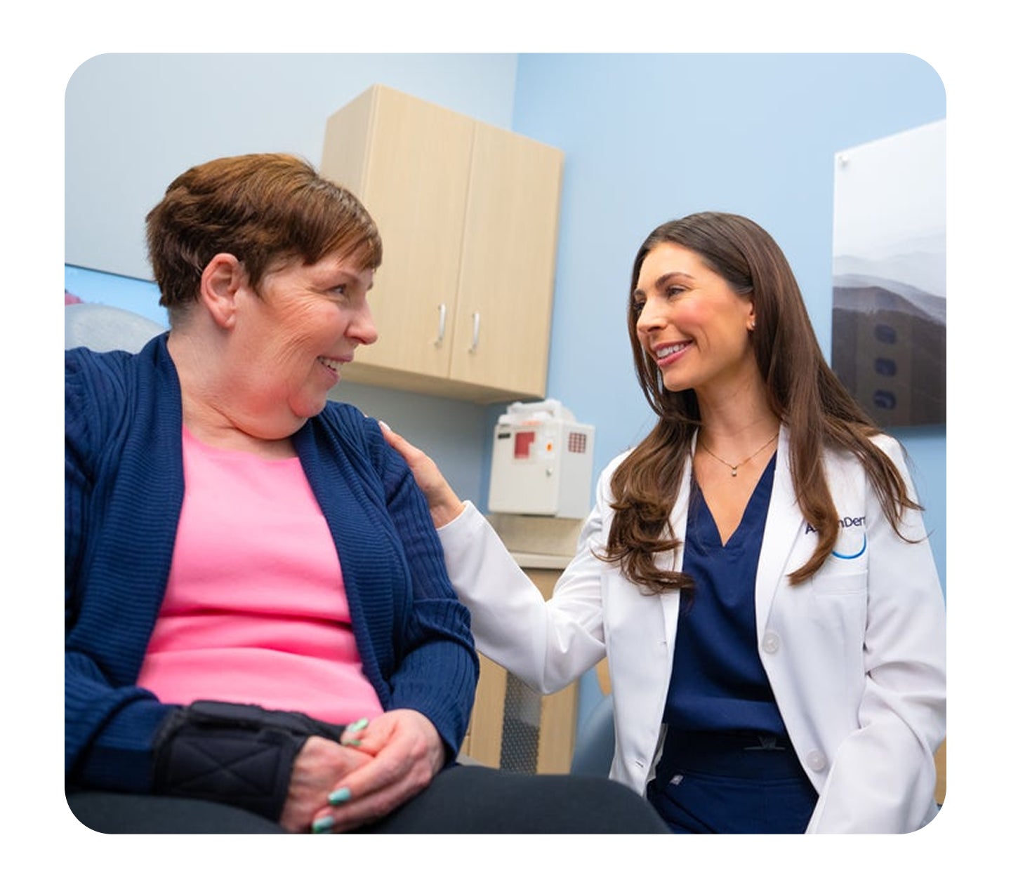 An Aspen Dental dentist in a white coat smiles while gently reassuring a seated female patient during a consultation in a dental exam room.
