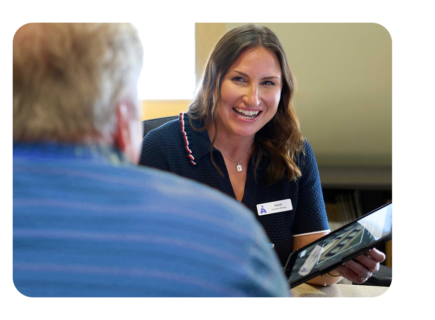 A friendly Aspen Dental team member, Susan, with long brown hair and wearing a navy polo with a name tag, smiles at a patient whose back is to the camera, while showing them in