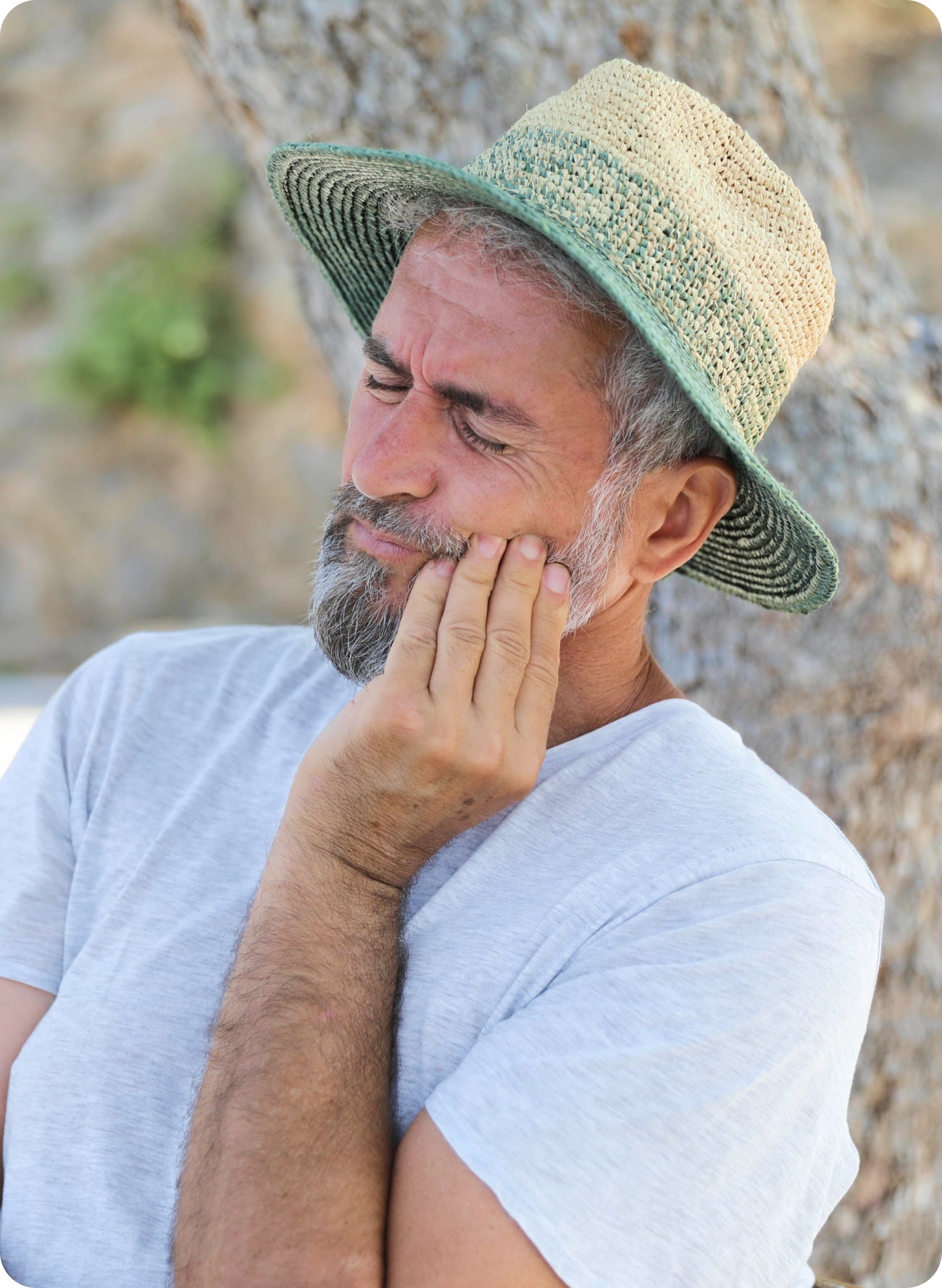 A bearded man in a straw hat, holding mouth due to pain