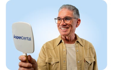 A smiling male patient wearing glasses admires his reflection in an Aspen Dental mirror against a light blue background.