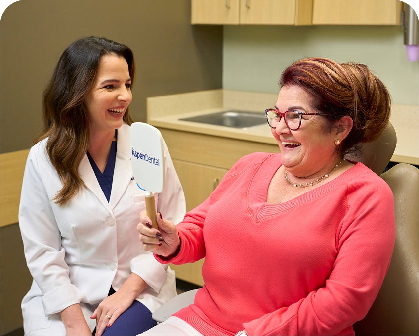 A happy female patient wearing glasses smiles while holding an Aspen Dental mirror as a dentist in a white coat smiles beside her during an appointment.