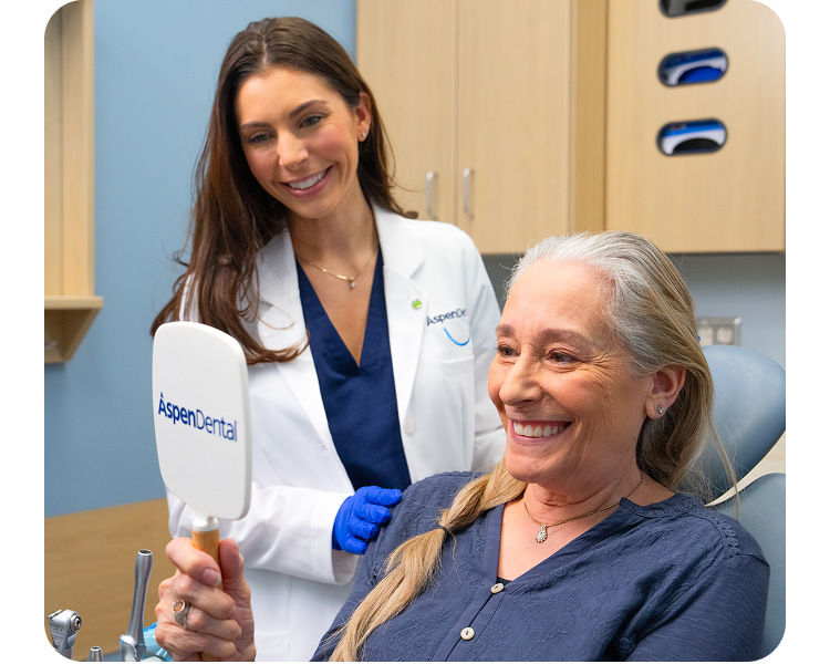 An older woman with long gray hair smiling brightly while looking at her reflection in a handheld mirror, while an Aspen Dental doctor stands smiling behind her.