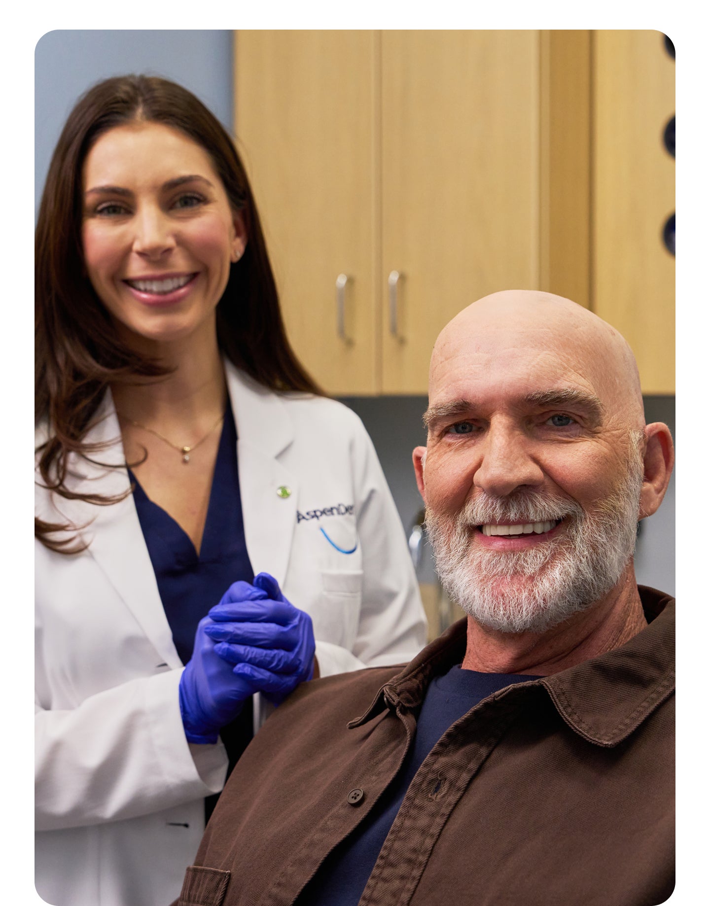 A smiling male patient sits in a dental exam chair while an Aspen Dental dentist in a white coat and blue gloves stands beside him, also smiling.
