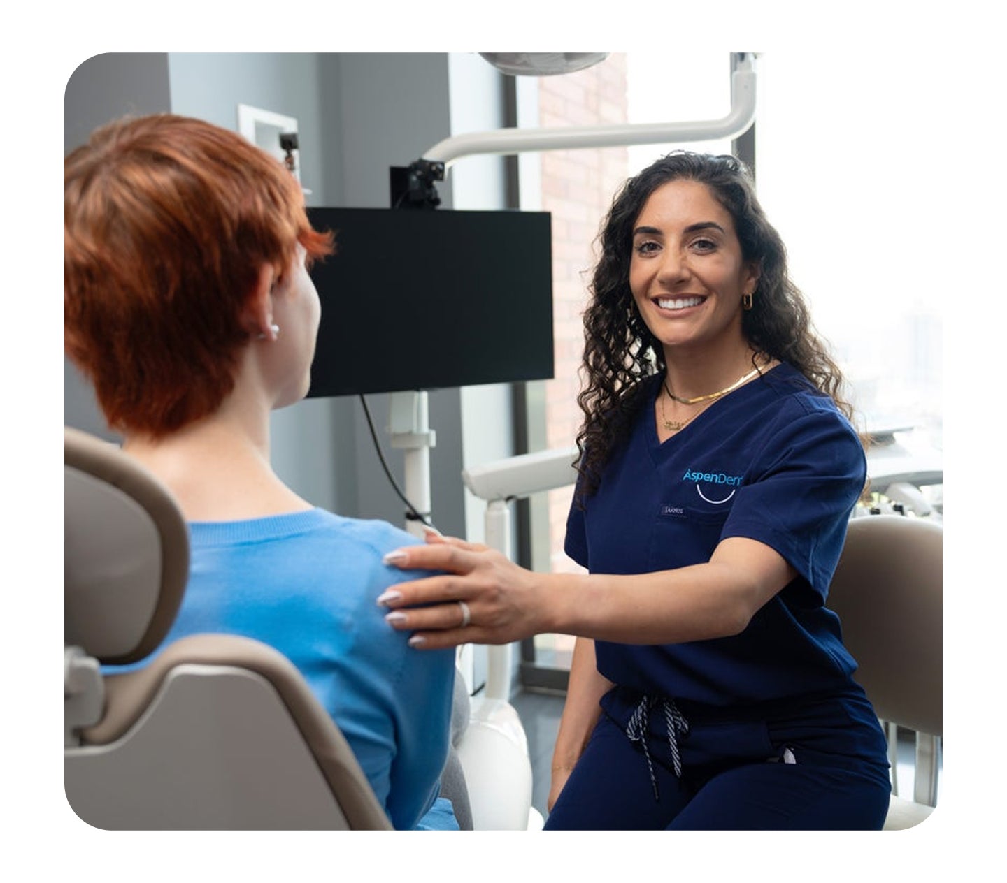 An Aspen Dental team member in navy scrubs smiles and gently places a hand on a patient’s shoulder while the patient sits in a dental chair, creating a calm and reassuring moment before treatment.