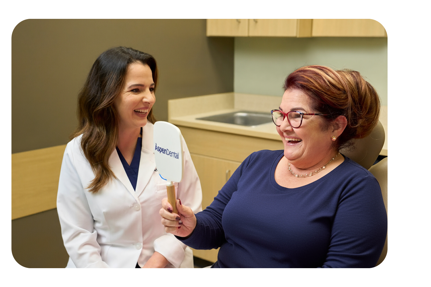 A smiling patient looks into an Aspen Dental mirror while a dentist in a white coat sits beside her, both sharing a joyful moment during a dental bridge consultation.