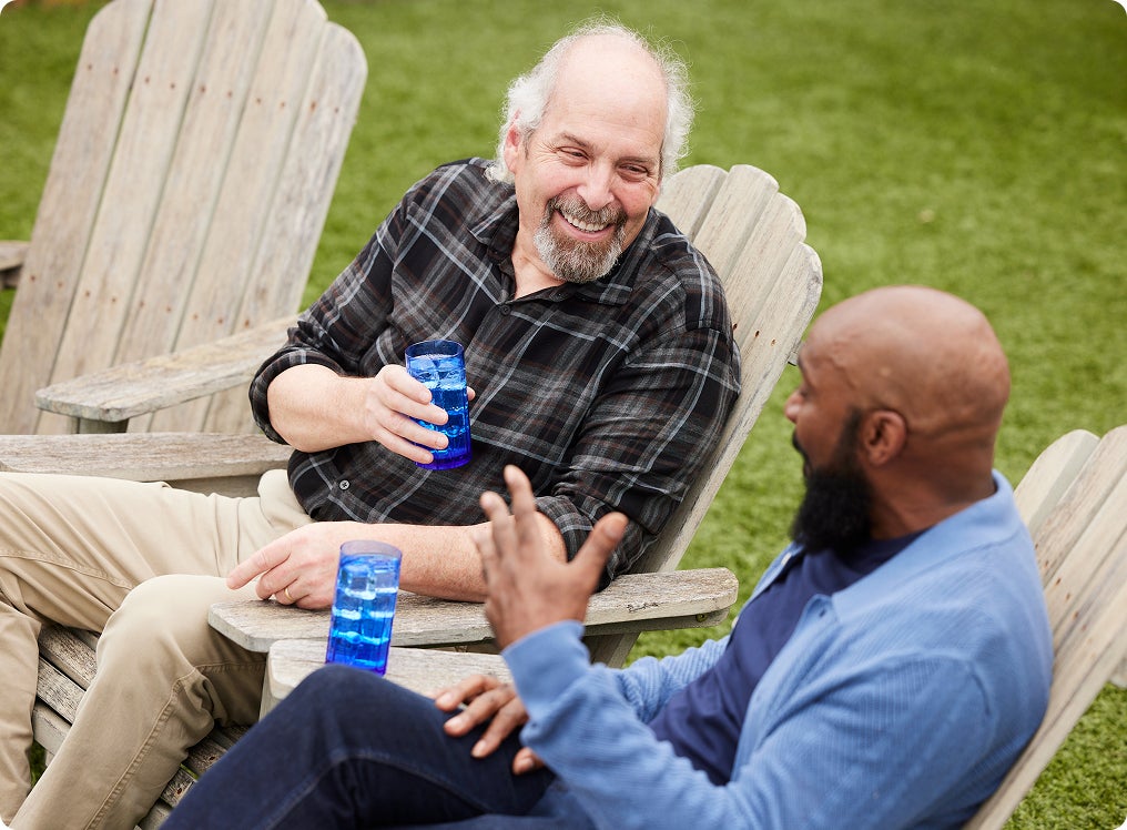 Two older men enjoying a relaxed conversation in wooden chairs on a lawn, reflecting confidence and comfort after receiving dental implants.